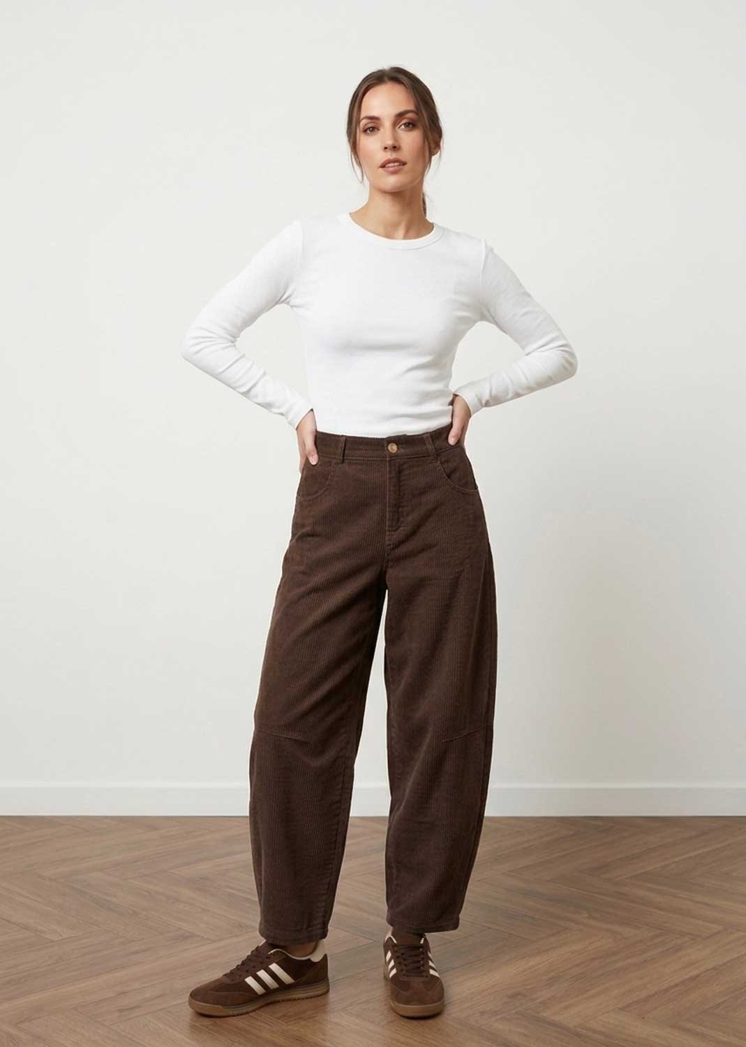 A woman stands in a studio wearing a fitted white long-sleeve top, dark brown wide-leg pants, and brown sneakers against a plain light wall and wood floor.