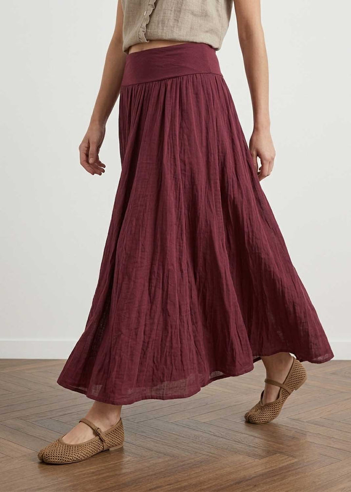 A woman wearing a high-waisted, flowing midi skirt in a deep burgundy or maroon crinkled fabric, paired with a beige crop top and woven brown flat shoes, standing on a wooden floor against a plain white wall.