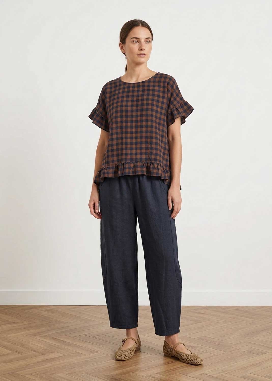 A woman stands in a studio wearing a dark brown and black checkered short-sleeve top with ruffled edges, dark navy wide-leg pants, and tan woven flats against a white wall and wood floor.