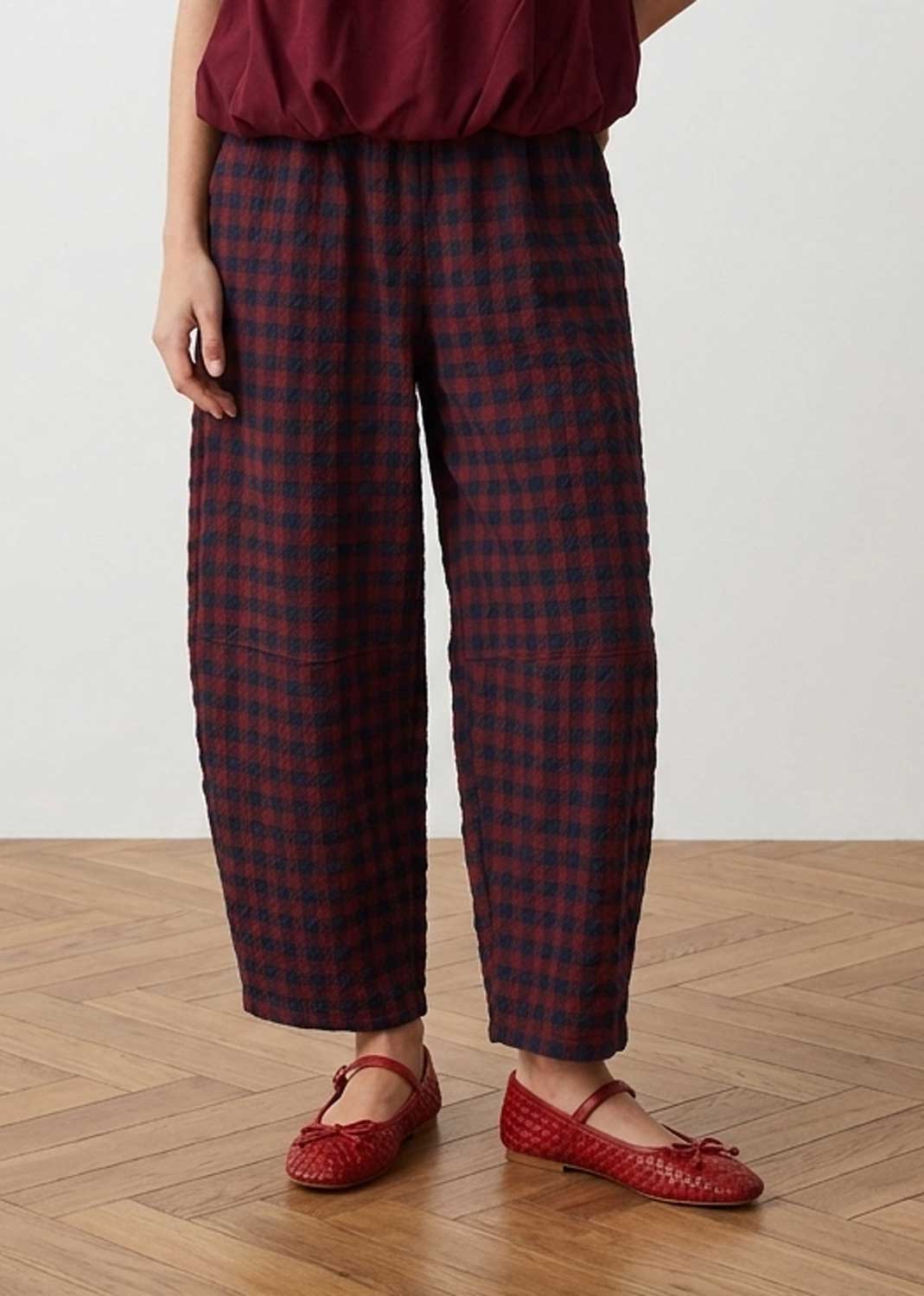 A person wearing loose cropped plaid pants in dark red and navy, paired with a burgundy top and red woven Mary Jane flats, standing on a light wood herringbone floor against a plain off-white wall.