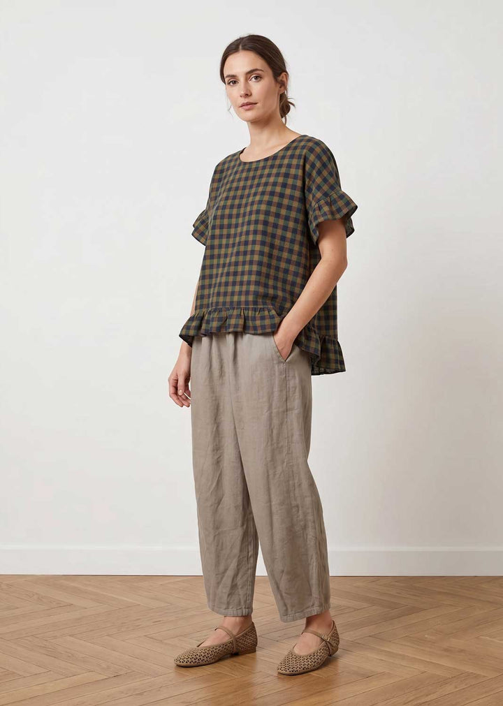 A woman modeling a short-sleeve plaid top in dark green and brown with a ruffled hem, paired with light beige linen trousers and brown knit flats, standing against a white wall on a wooden floor.