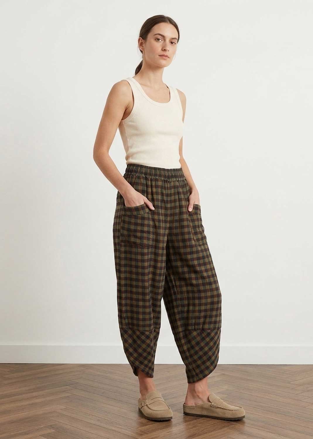A woman modeling loose, baggy brown and khaki plaid trousers with a cream ribbed tank top and beige slip-on loafers, standing against a white wall on a wood floor.
