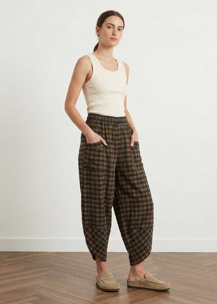 A woman modeling loose, baggy brown and khaki plaid trousers with a cream ribbed tank top and beige slip-on loafers, standing against a white wall on a wood floor.