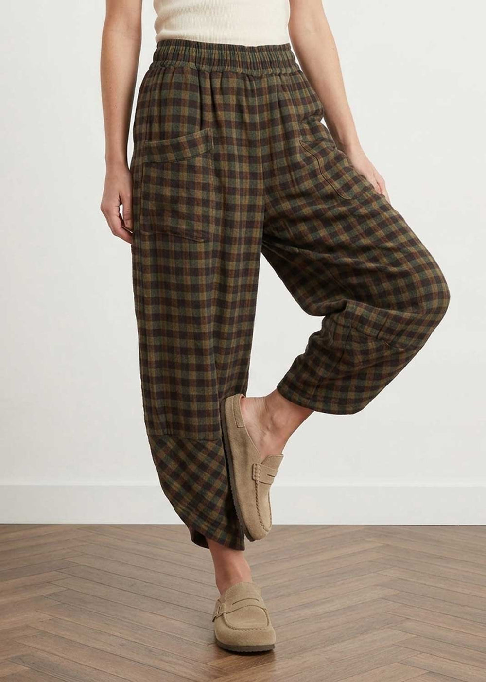A person wearing loose, high-waisted plaid pants in dark green and brown tones, paired with beige slip-on loafers, standing on a wood floor against a plain light wall.