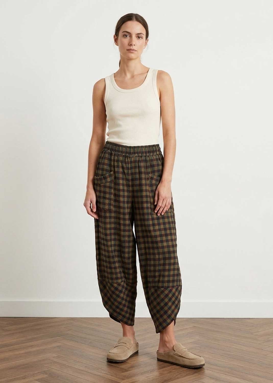 A woman stands in a studio wearing a cream ribbed tank top, dark brown and black plaid cropped pants, and beige slip-on shoes against a white wall and wood floor.