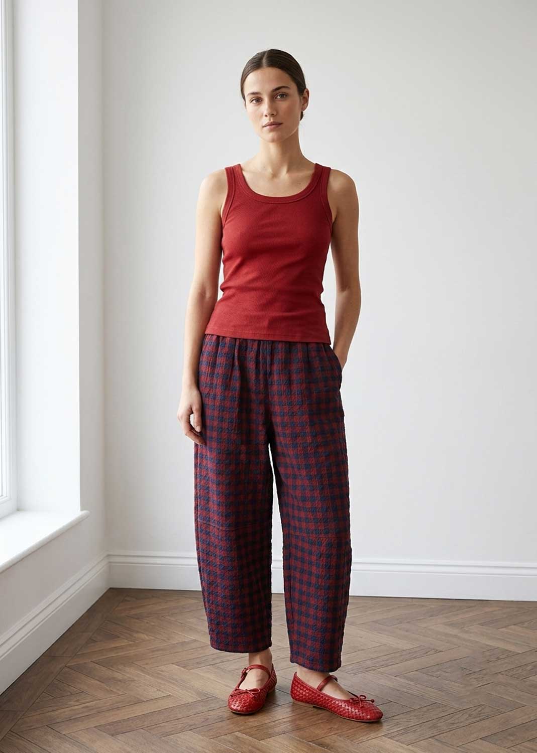 A woman stands in a bright room with white walls and a wood floor, wearing a sleeveless red top and loose dark plaid pants with red shoes.