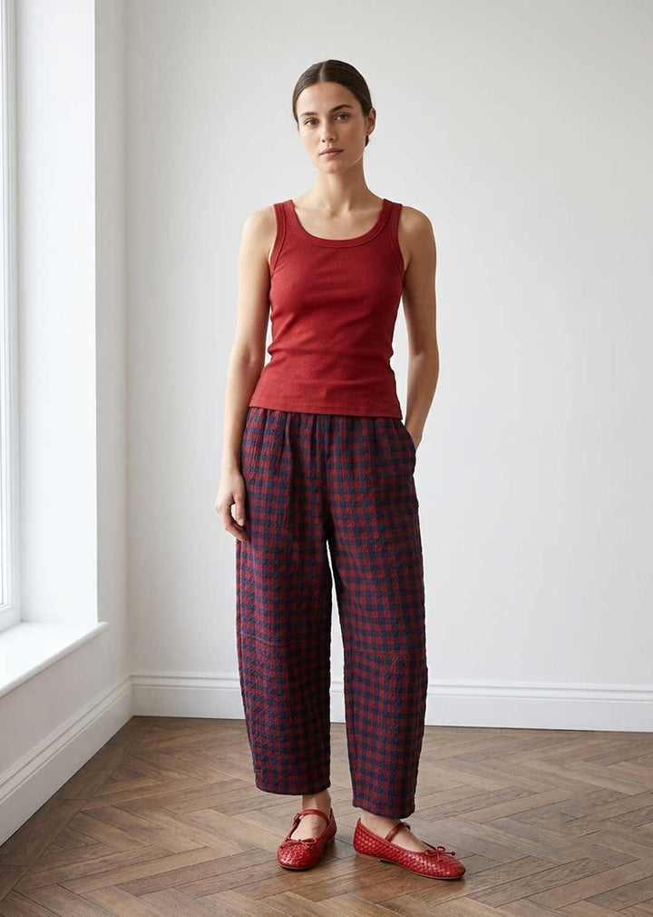 A woman stands in a bright room with white walls and a wood floor, wearing a sleeveless red top and loose dark plaid pants with red shoes.