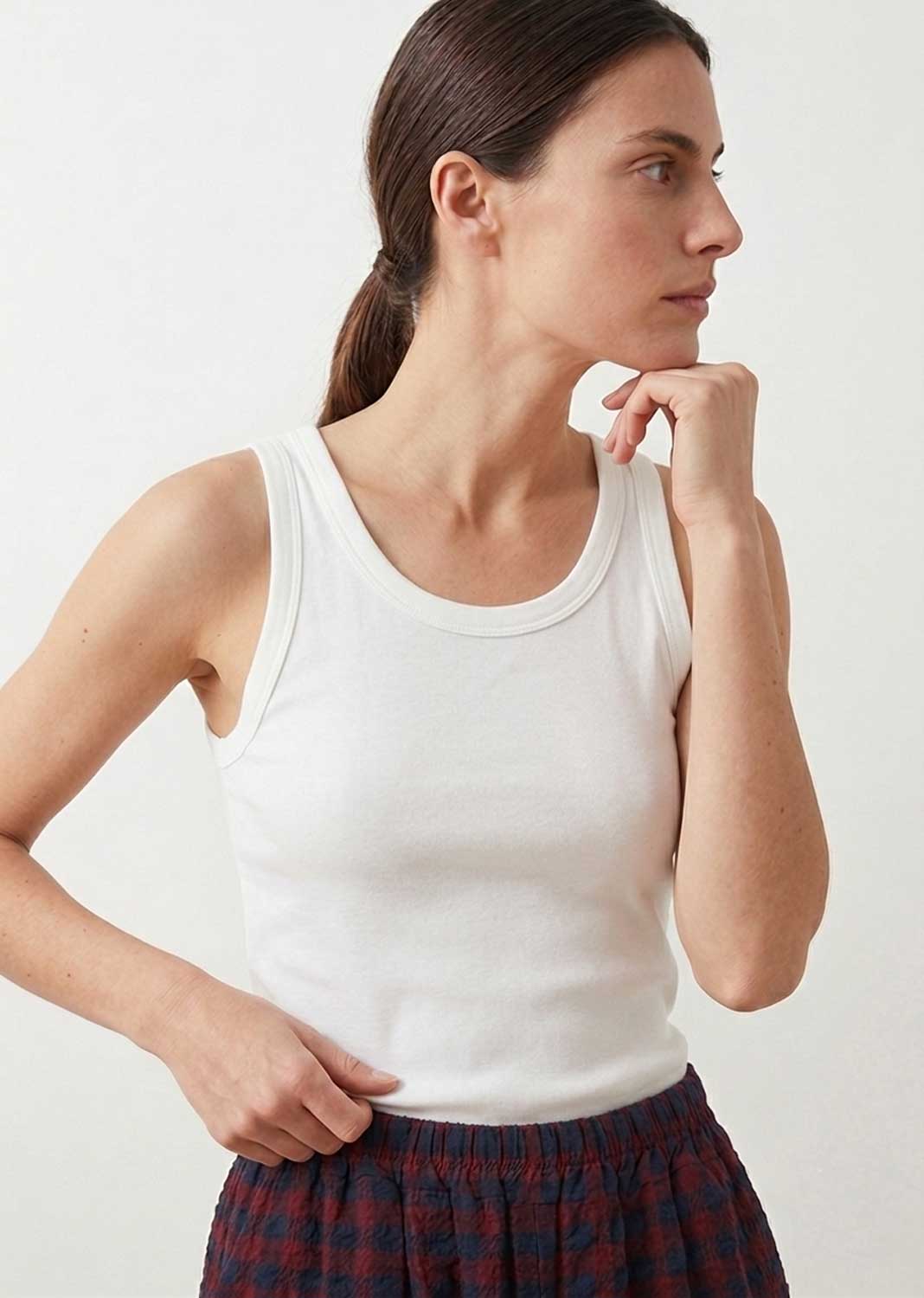 A woman wearing a white ribbed tank top and a red and navy plaid skirt, posing against a plain white studio background.