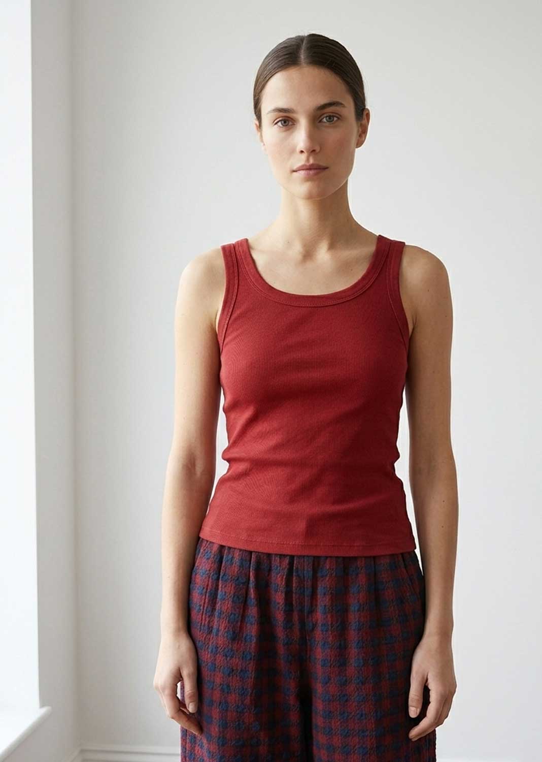 A woman modeling a sleeveless red tank top paired with dark red and navy plaid pants against a plain white wall.
