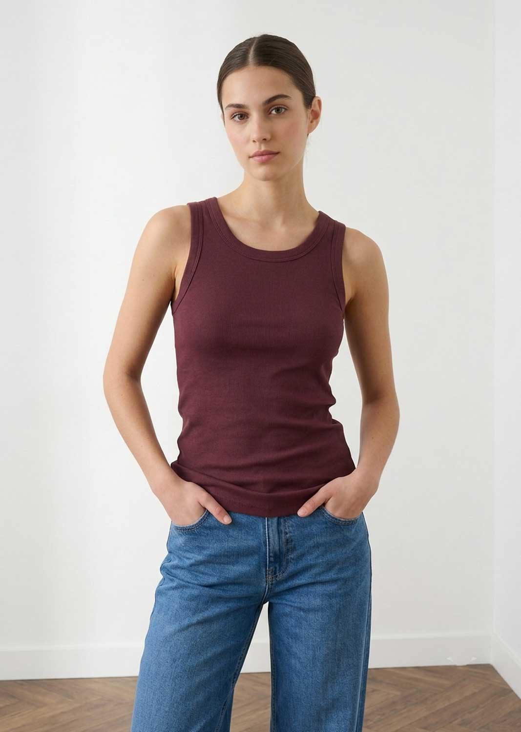 A woman modeling a sleeveless crew-neck tank top in deep burgundy, paired with blue jeans, standing against a plain white wall.