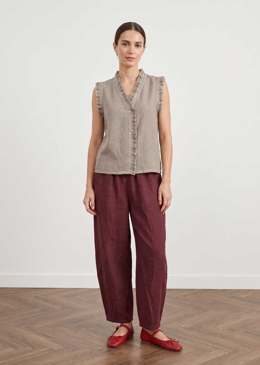 A woman stands in a studio wearing a sleeveless taupe linen vest with ruffle trim and dark burgundy linen trousers, paired with red flats, against a white wall and wood floor.