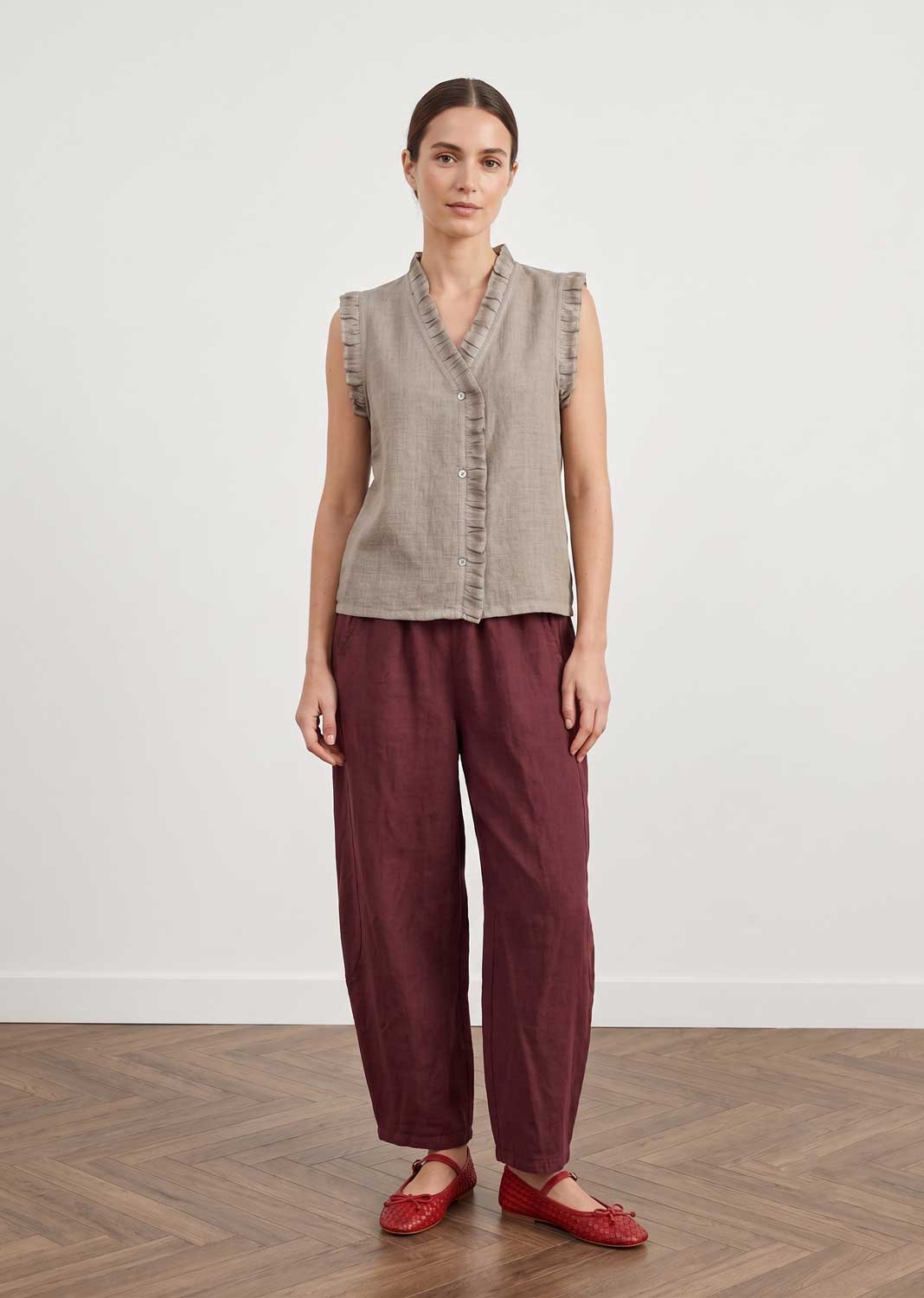 A woman stands in a studio wearing a sleeveless taupe linen vest with ruffle trim and dark burgundy linen trousers, paired with red flats, against a white wall and wood floor.