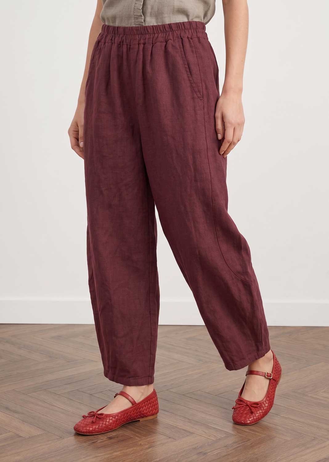 A person wearing loose, wide-leg trousers in a deep burgundy or maroon color, paired with red Mary Jane flats, standing on a wooden parquet floor against a plain white wall.