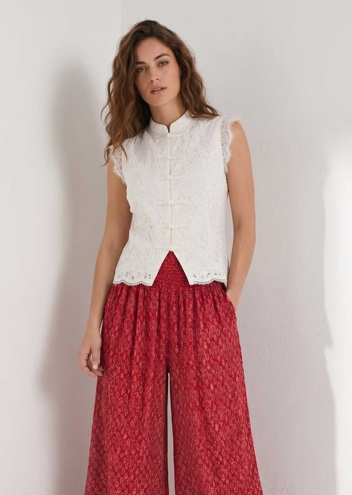 A woman modeling a sleeveless white lace vest with a mandarin collar over a red patterned wide-leg pants, standing against a plain light gray wall.