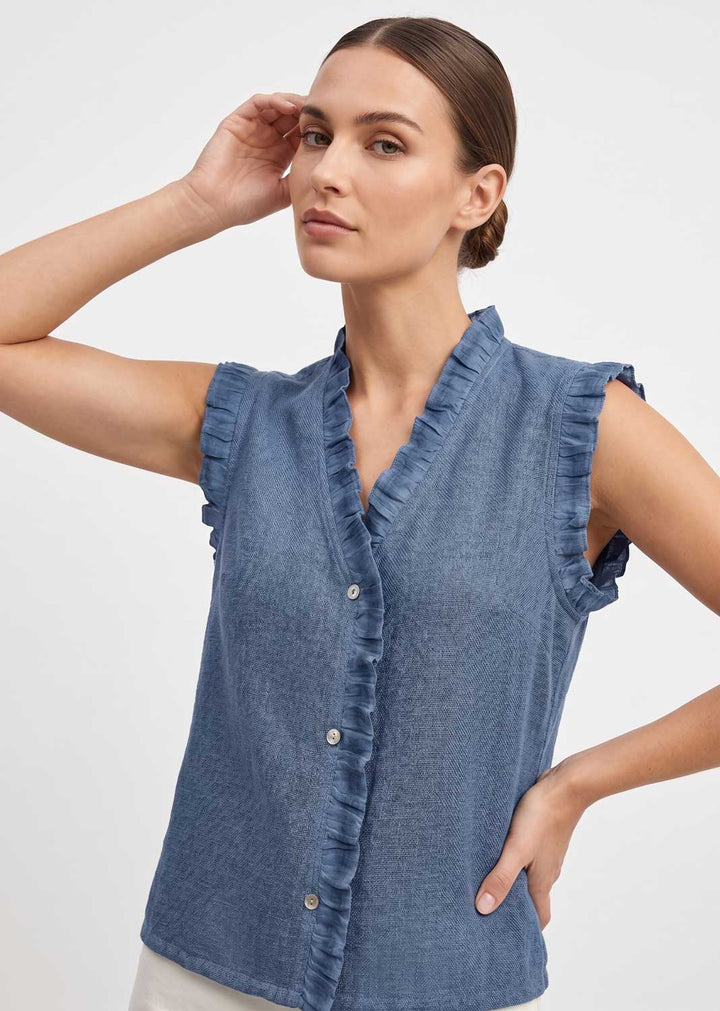 A woman wearing a sleeveless blue chambray button-front top with ruffle trim, photographed against a clean light gray studio background.