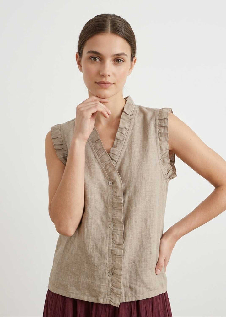 A woman modeling a sleeveless beige linen blouse with ruffle trim and a button front, paired with a dark burgundy skirt, against a plain light gray studio background.