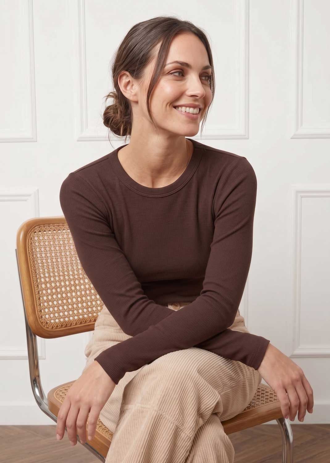 A woman with her hair in a bun sits on a wooden cane chair, wearing a dark brown long-sleeve crewneck top and beige ribbed trousers, set against a white paneled wall.