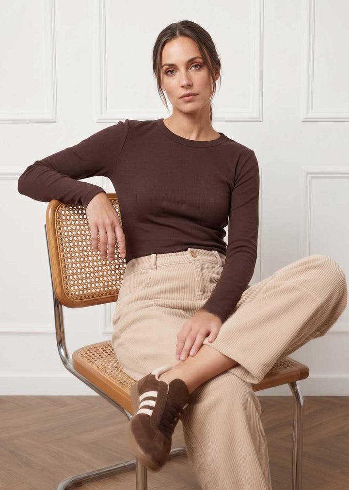 A woman sits on a wooden chair with a woven back, wearing a dark brown long-sleeve crewneck top, beige corduroy trousers, and brown suede shoes with white stripes, set against a white paneled wall.