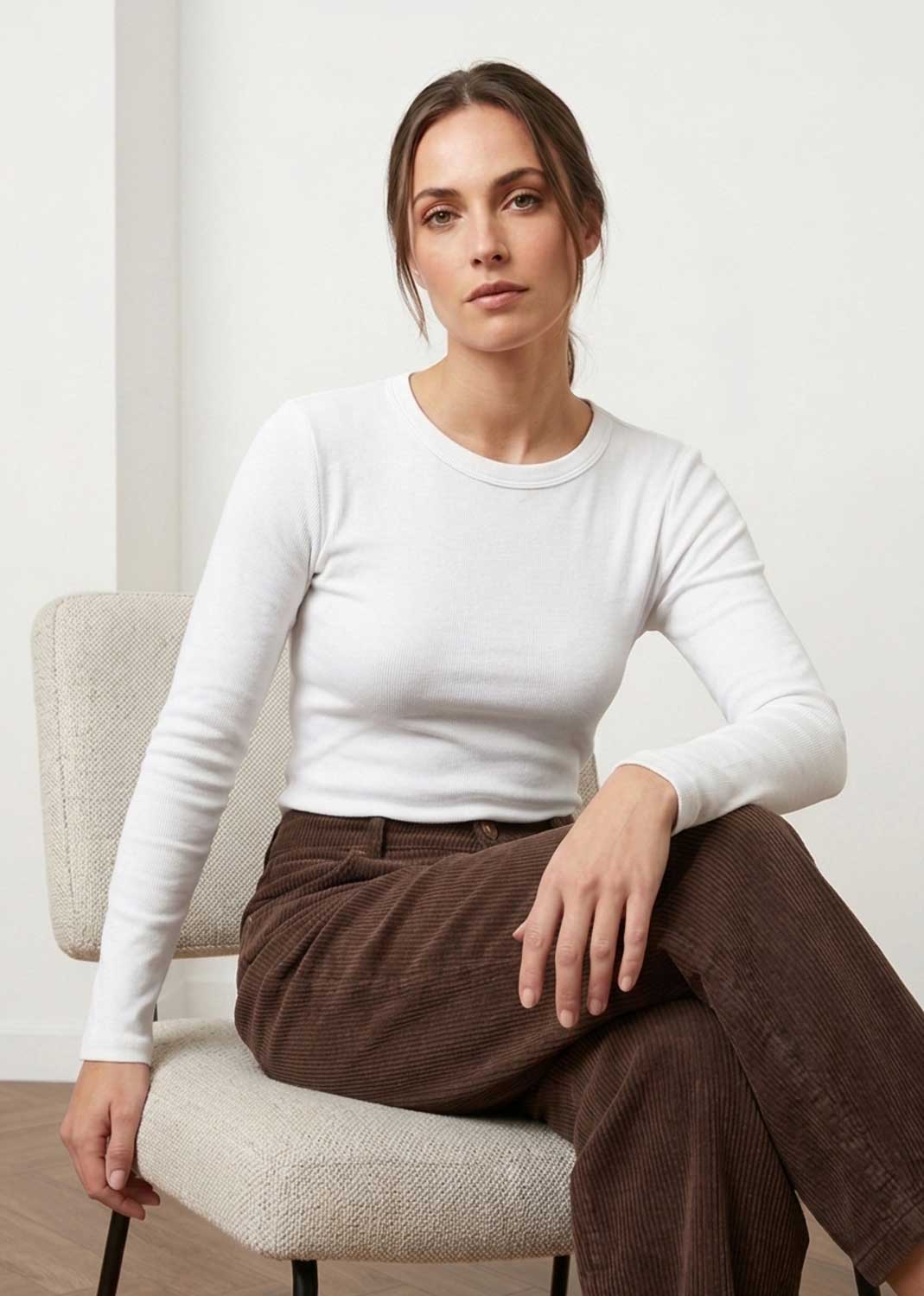 A woman sits on a light beige upholstered chair wearing a fitted white long-sleeve crewneck top and brown corduroy pants in a bright indoor setting.