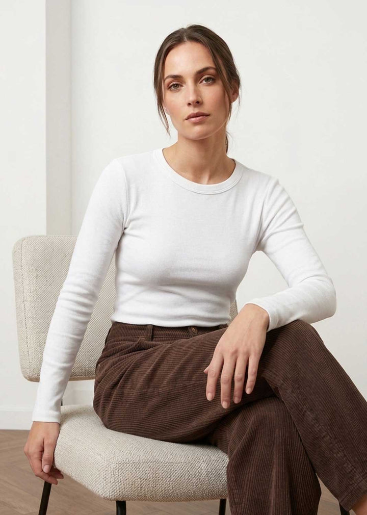 A woman sits on a light beige upholstered chair wearing a fitted white long-sleeve crewneck top and brown corduroy pants in a bright indoor setting.