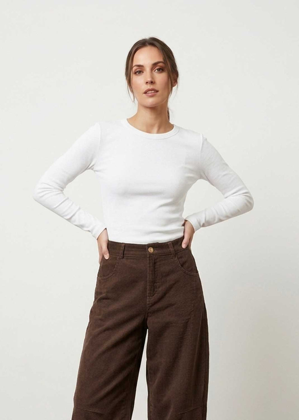 A woman modeling a white long-sleeve crewneck top paired with brown wide-leg pants, standing against a plain light gray studio background.