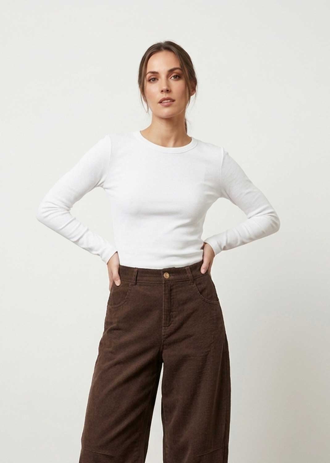A woman modeling a white long-sleeve crewneck top paired with brown wide-leg pants, standing against a plain light gray studio background.