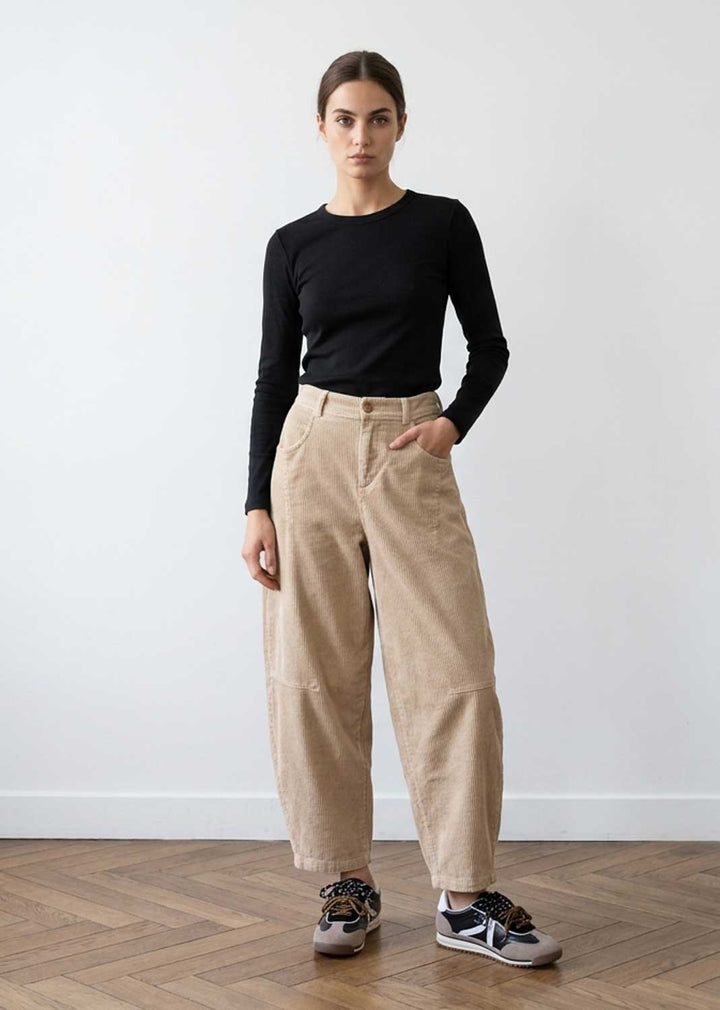 A woman stands in a studio wearing a fitted black long-sleeve top, loose beige corduroy pants, and gray sneakers against a plain white wall and wood floor.