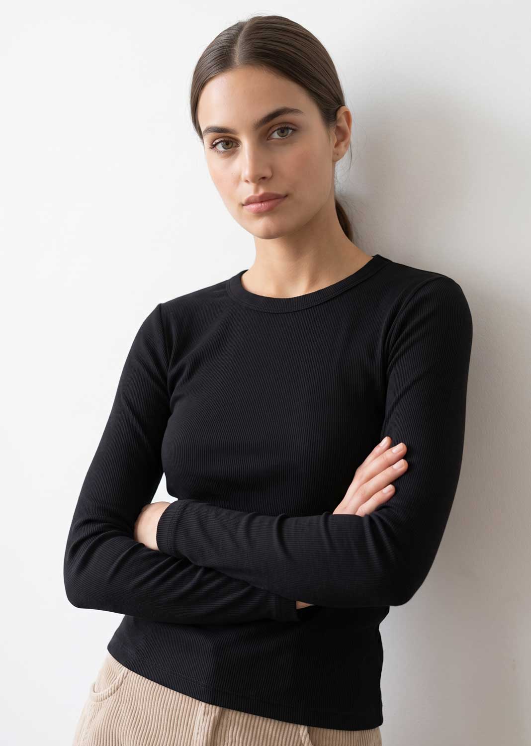 A woman with her hair pulled back wears a fitted black long-sleeve crewneck top, standing with arms crossed against a plain light gray wall.
