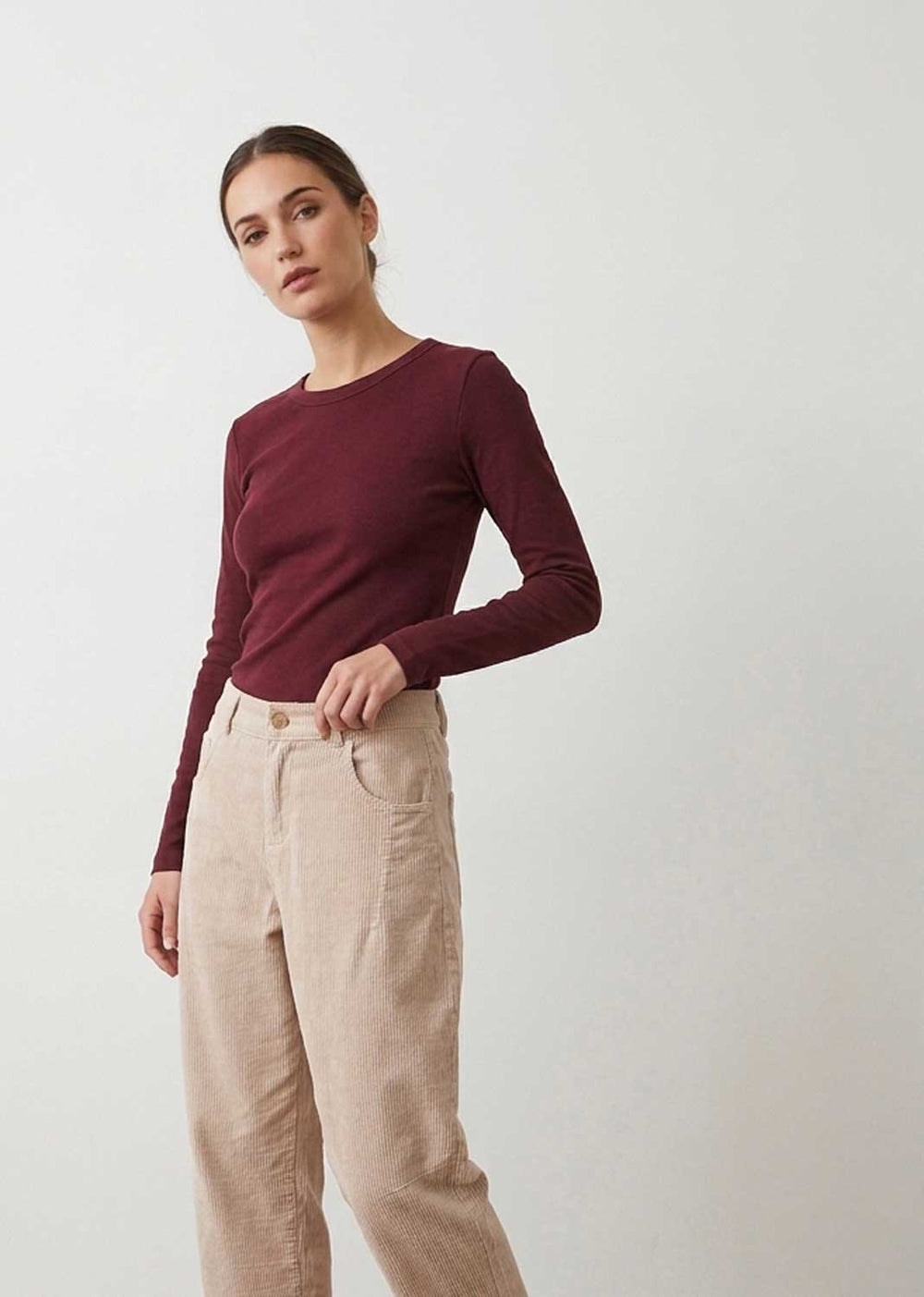 A woman modeling a long-sleeve crewneck top in deep burgundy paired with light beige linen trousers, set against a plain light gray studio background.