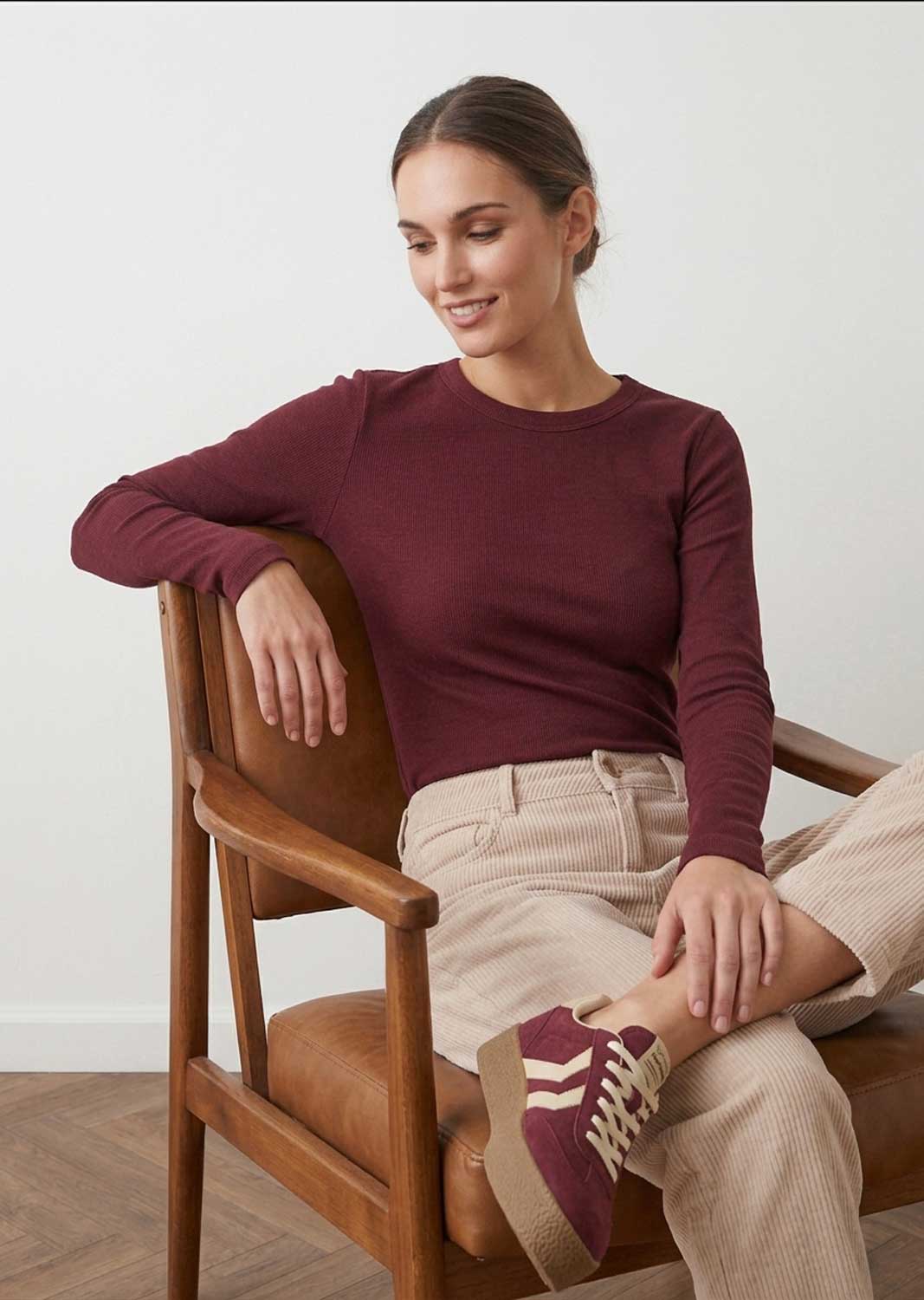 A woman sitting on a wooden chair wearing a long-sleeve crewneck knit top in burgundy, beige corduroy trousers, and burgundy sneakers with white stripes.