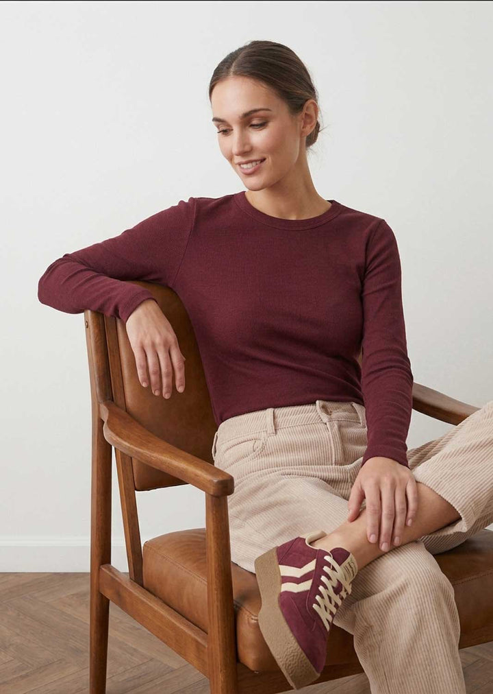 A woman sitting on a wooden chair wearing a long-sleeve crewneck knit top in burgundy, beige corduroy trousers, and burgundy sneakers with white stripes.