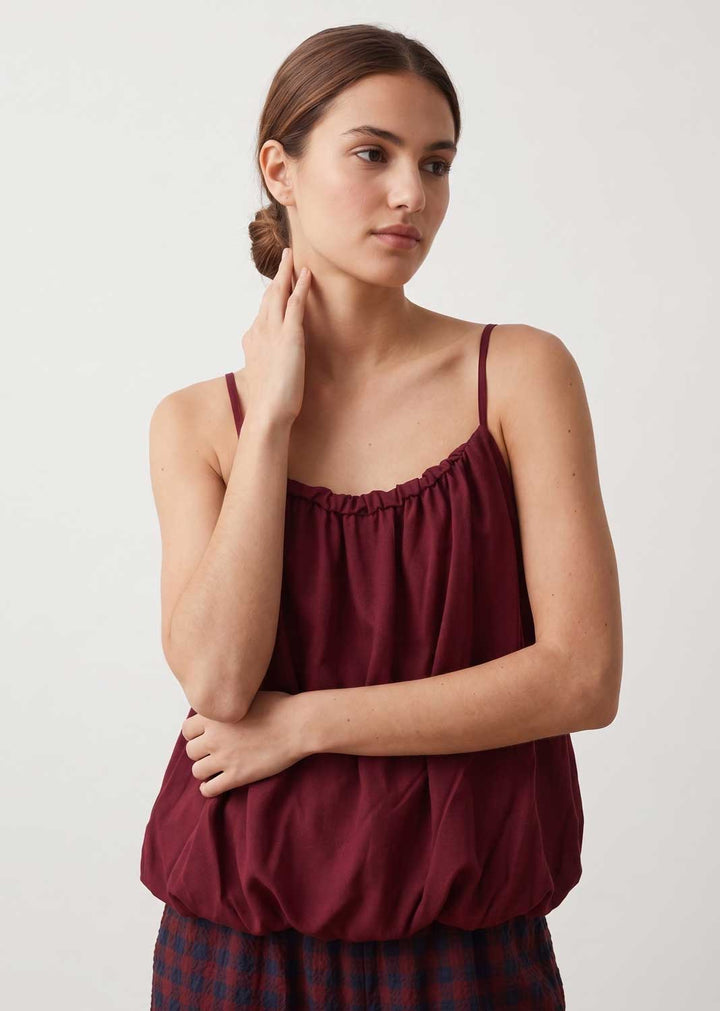 A woman wearing a burgundy spaghetti-strap camisole top and a dark plaid skirt, posing against a light gray studio background.