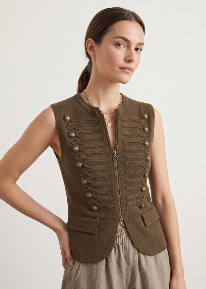 A woman modeling a sleeveless olive-brown military-style vest with decorative button details and a front zipper, paired with light beige trousers, against a plain light gray background.