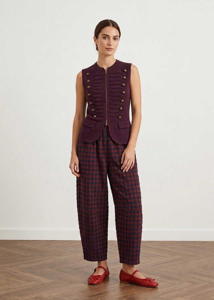 A model stands in a studio wearing a sleeveless deep purple vest with gold-tone buttons and matching dark red and purple plaid cropped trousers, paired with red Mary Jane flats.