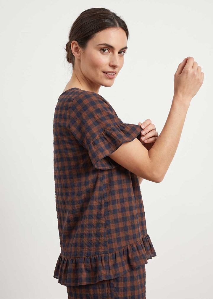 A woman modeling a brown and navy blue plaid short-sleeve top with ruffled trim, shown from a side-back angle against a plain light gray background.