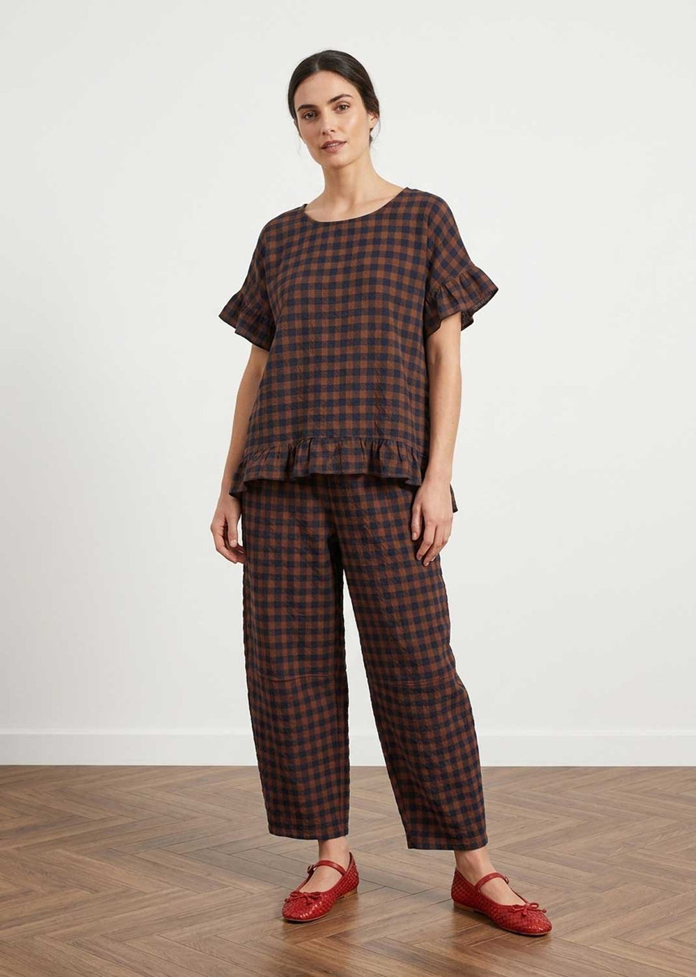 A woman modeling a matching two-piece plaid lounge set consisting of a short-sleeve top with ruffled sleeves and wide-leg cropped pants, in a dark brown and black checkered pattern, paired with red flat shoes against a white wall and wood floor.