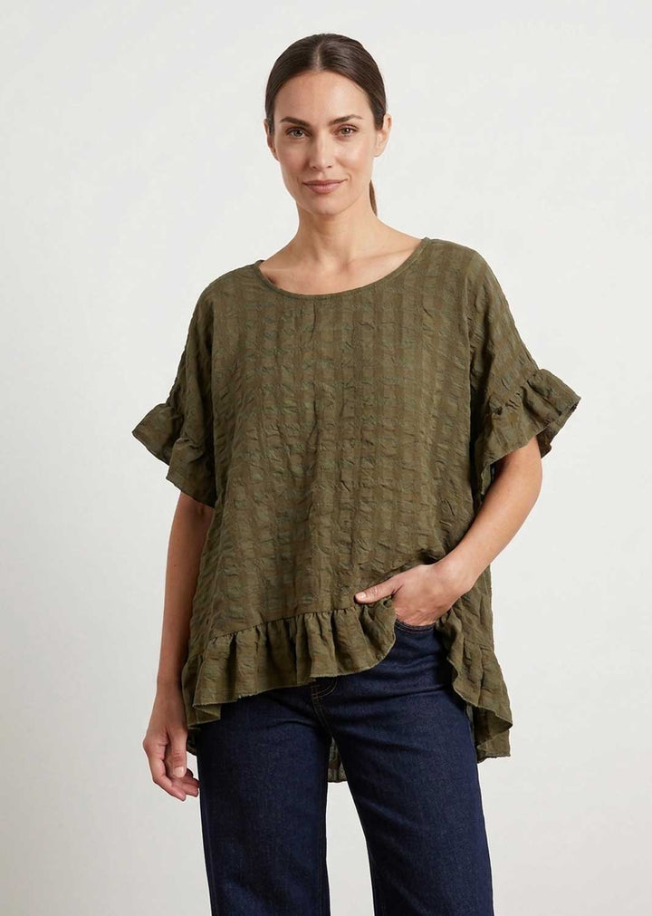 A woman modeling a textured seersuker olive green short-sleeve top with ruffled sleeves and hem, paired with dark blue jeans, against a plain light gray background.