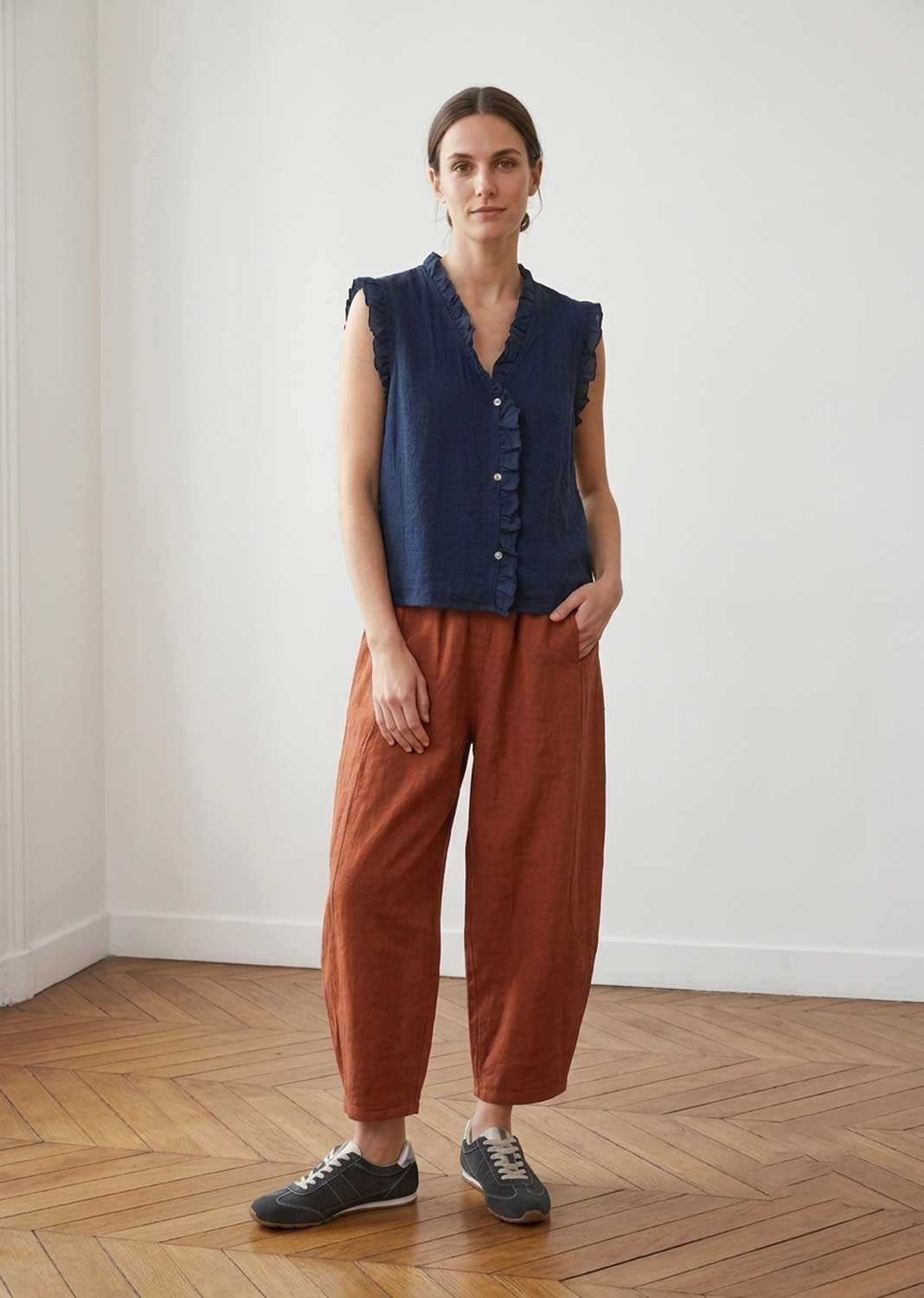 A woman stands in a bright room with a wood floor and white walls, wearing a sleeveless navy blue button-front top and rust-brown wide-leg pants with dark gray sneakers.