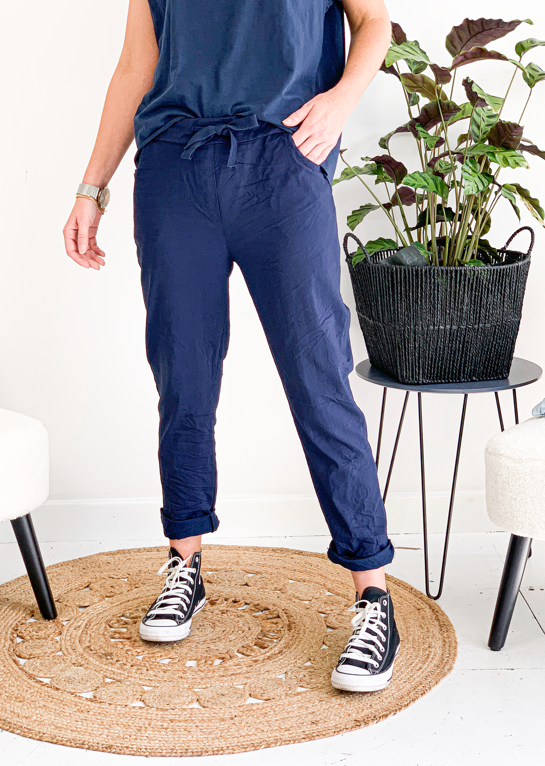 A person standing in an indoor setting wearing navy magic pants with a drawstring waist, side pockets, and roll-up leg. They are paired with a white top and beige flat shoes, carrying a white tote bag.